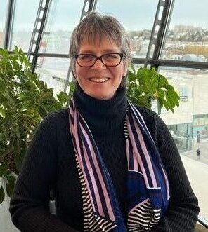 Eine lächelnde Frau mit kurzen grauen Haaren und Brille trägt einen schwarzen Rollkragenpullover und einen bunt gestreiften Schal. Sie sitzt im Innenbereich vor großen Fenstern, von denen aus Pflanzen zu sehen sind. Draußen ist eine Stadtlandschaft zu sehen.